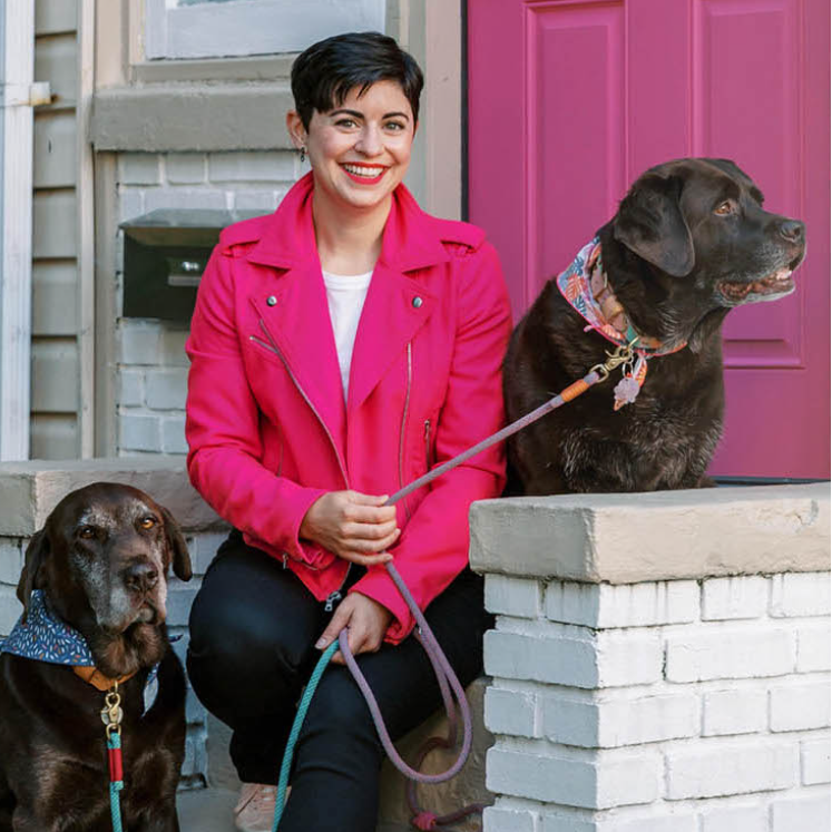 A woman in a bright pink jacket sits on a white brick porch with two brown dogs, one sitting and one standing, in front of a pink door.